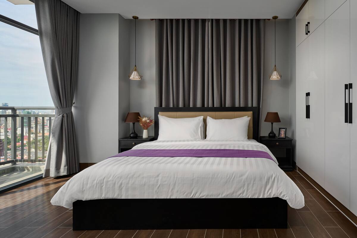 A modern hotel room with a large bed, two bedside tables with lamps, a view of the city through floor-to-ceiling windows, and a balcony.