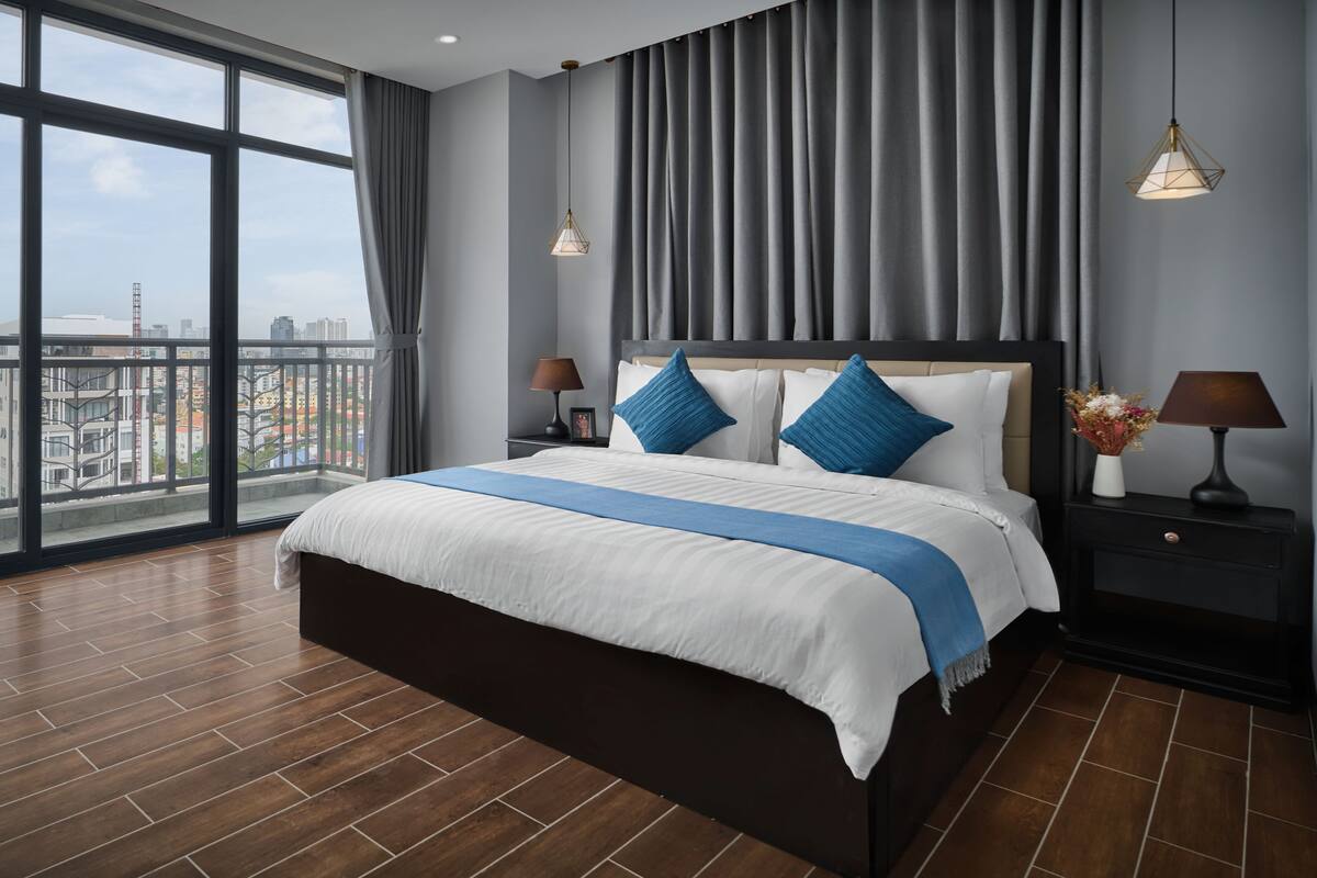 A modern hotel room with a large bed, a nightstand, a lamp, and a view of the city through large windows.
