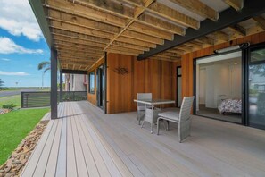 Uma área externa coberta com deck de madeira, uma mesa com cadeiras e uma porta de vidro que dá para um quarto.