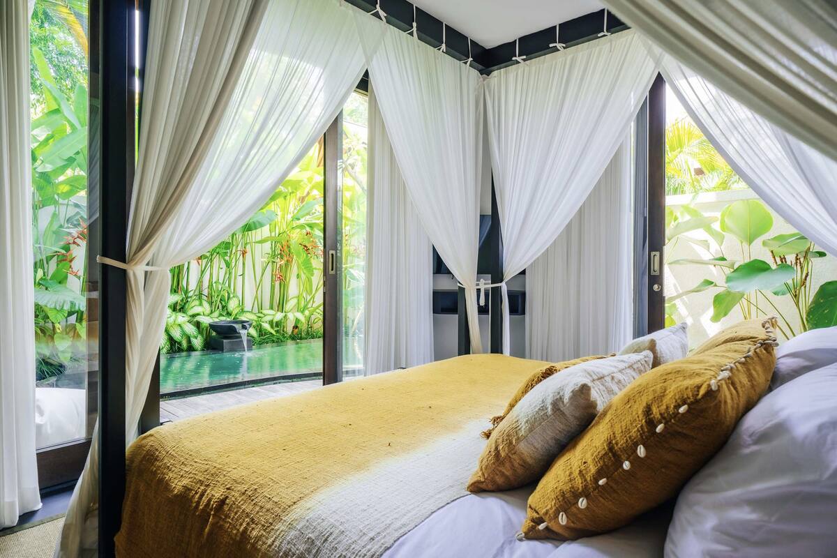 A bedroom with a large bed, white curtains, and a view of a lush garden and pool.