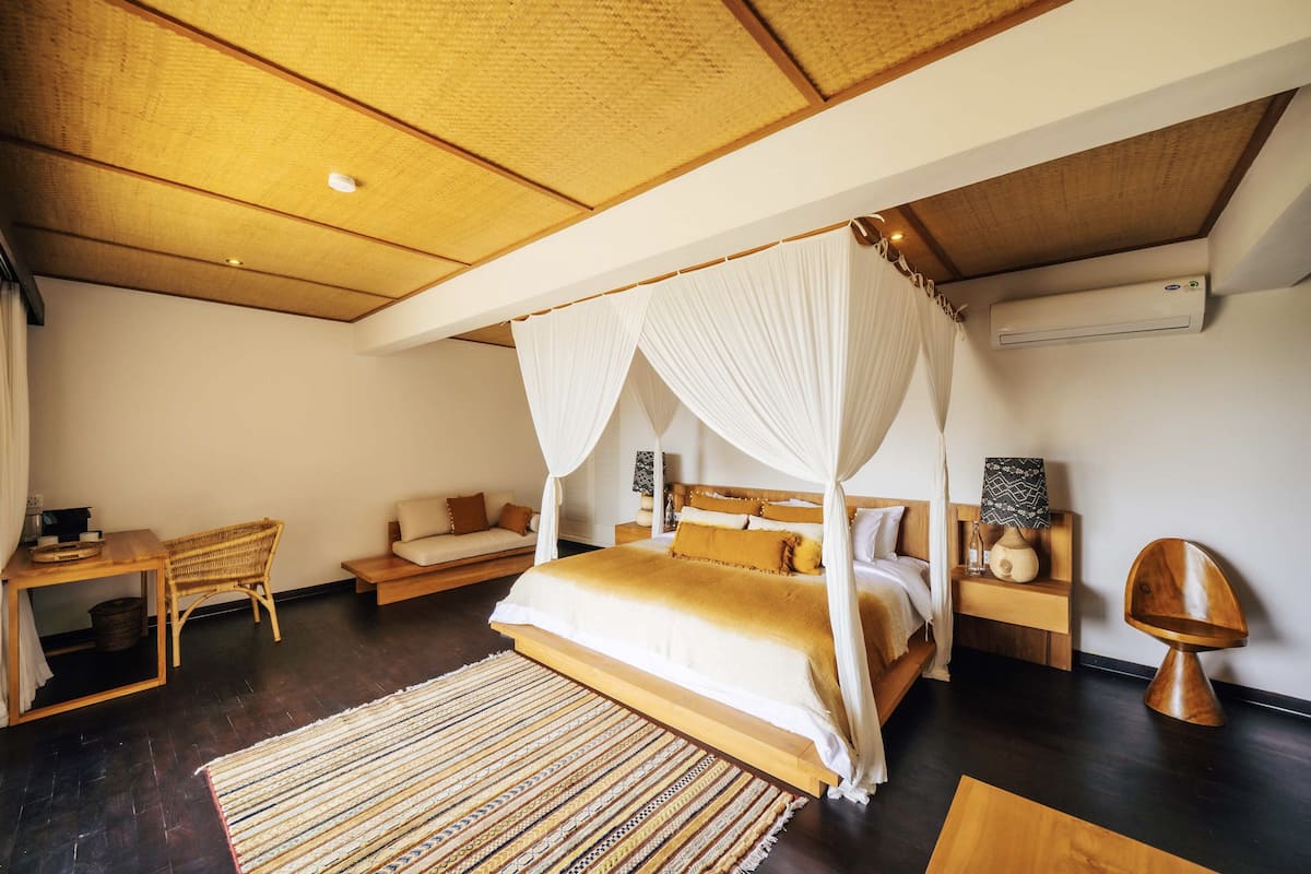 A bedroom with a four-poster bed, a wooden headboard, a bedside table with a lamp, a chair, and a small table with a basket.