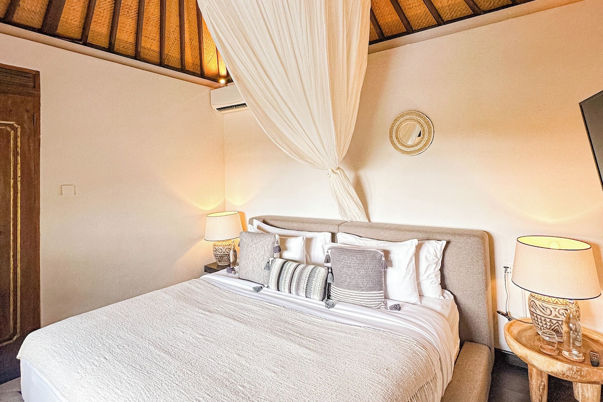 A hotel room with a large bed, a canopy, two bedside tables with lamps, and a wall clock.