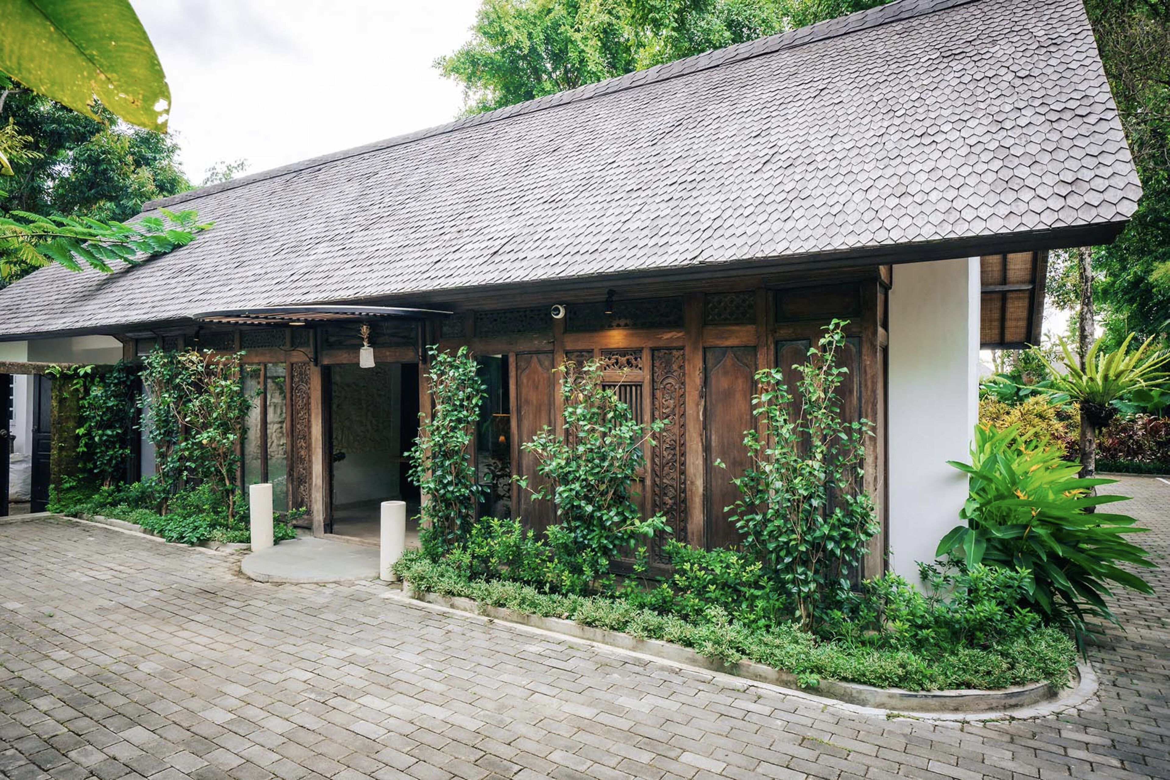 A traditional wooden building with a tiled roof, surrounded by lush greenery and a paved entrance.