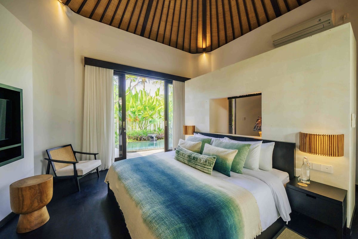 A modern hotel room with a large bed, a chair, a small table, and a balcony with a view of greenery.