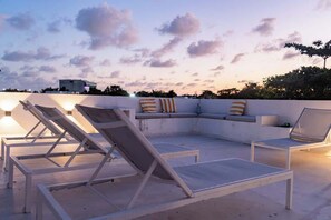 Un área de lounge en la azotea con sillones y un sofá, una pared blanca y un cielo al atardecer.