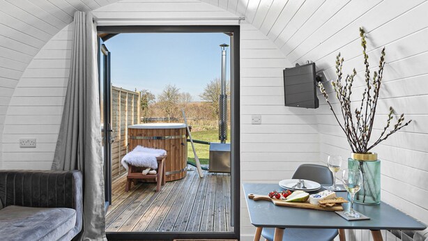 A cozy interior with a wooden hot tub, a dining table set with food and drinks, and a view of the outdoors. - Bramley Glamping Pod- Free Wi-Fi, Hot Tub (Wells)