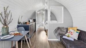 A compact, modern interior with a dining area, kitchenette, and a cozy seating area with a sofa and cushions. - Bramley Glamping Pod- Free Wi-Fi, Hot Tub (Wells)