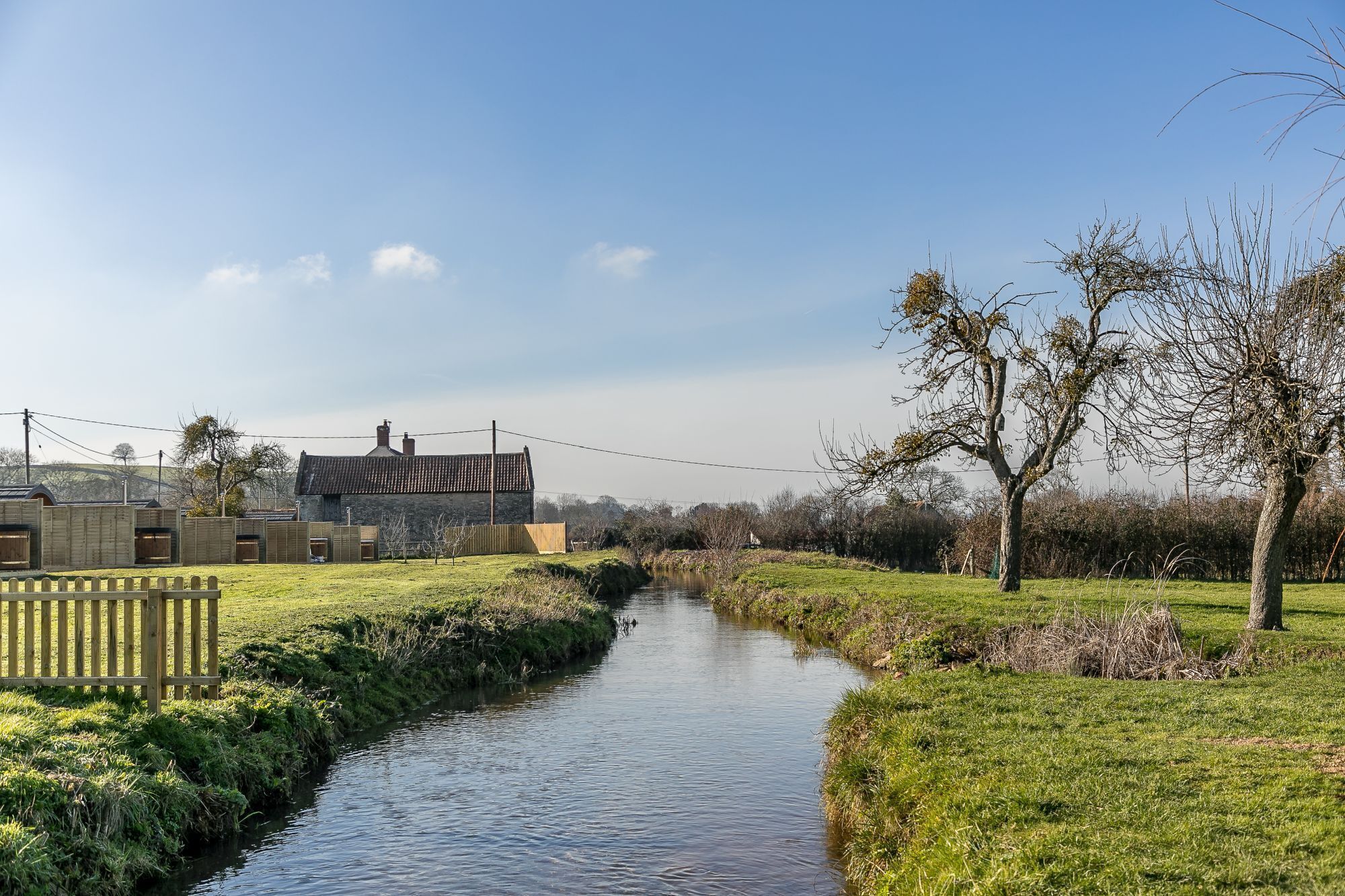 A rural landscape with a narrow water channel, a stone building with a tiled roof, and a wooden fence.
