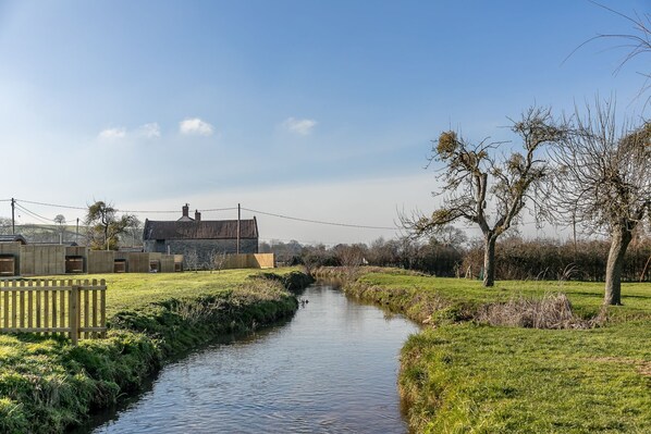 A rural landscape with a narrow water channel, a stone building with a tiled roof, and a wooden fence. - Bramley Glamping Pod- Free Wi-Fi, Hot Tub (Wells)