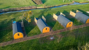 A row of wooden cabins with grey roofs, arranged in a linear fashion along a gravel path, surrounded by lush green fields and a small water body. - Windsor Glamping Pod- Free Wi-Fi, Hot Tub (Wells)