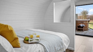 A cozy bedroom with a bed, a wooden side table with a tray of fruit and a book, and a view of an outdoor deck. - Morgan Sweet Glamping Pod - Free Wi-Fi (Wells)
