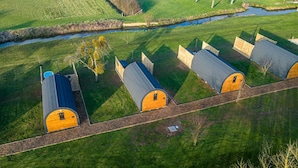 A row of wooden cabins with grey roofs, arranged in a linear fashion along a gravel path, surrounded by lush green fields and a small water body. - Morgan Sweet Glamping Pod - Free Wi-Fi (Wells)