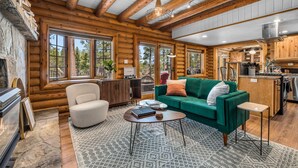 A log cabin interior with a green sofa, a white armchair, a coffee table, and a kitchen area with wooden cabinets and a stove.