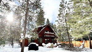 A log cabin with a balcony, surrounded by snow-covered ground and tall pine trees.