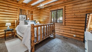 A log cabin bedroom with a wooden bed, a nightstand, a lamp, a framed picture, and a window with a view of trees.