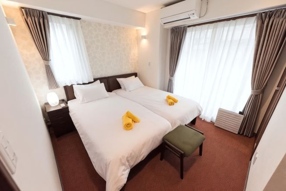 A hotel room with two beds, a nightstand, a lamp, a bench, and curtains.