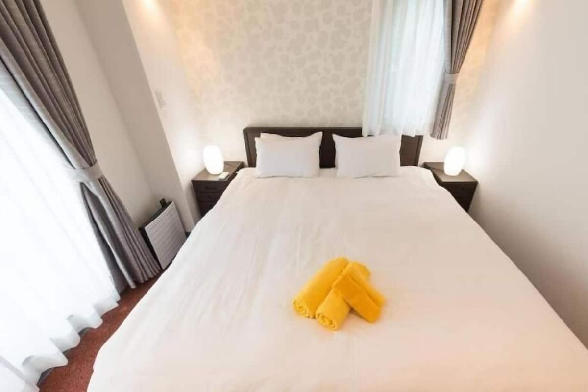 A hotel room with a large bed, white linens, two bedside tables with lamps, and a window with curtains.