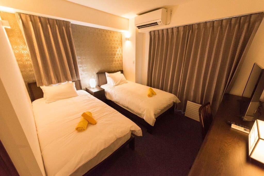 A hotel room with two beds, a desk, a TV, and an air conditioning unit.