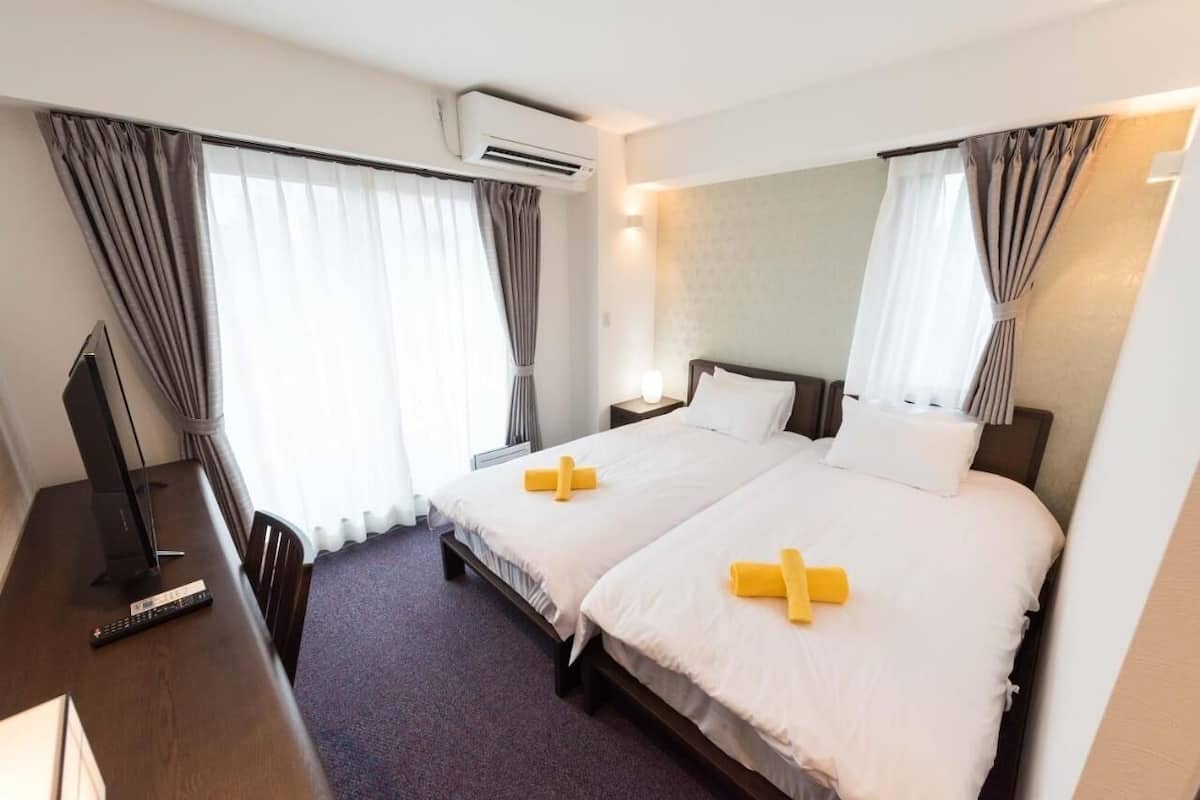 A hotel room with two beds, a desk with a TV, and an air conditioning unit.