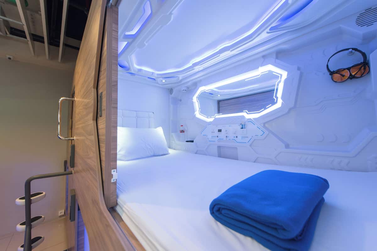 A modern, well-lit room with a neatly made bed, a control panel, and a pair of headphones on the wall.