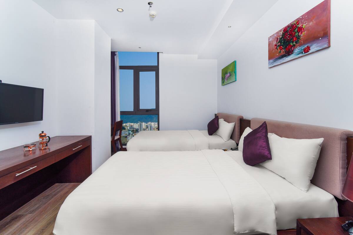 A hotel room with two beds, a TV, a desk, and a window with a city view.