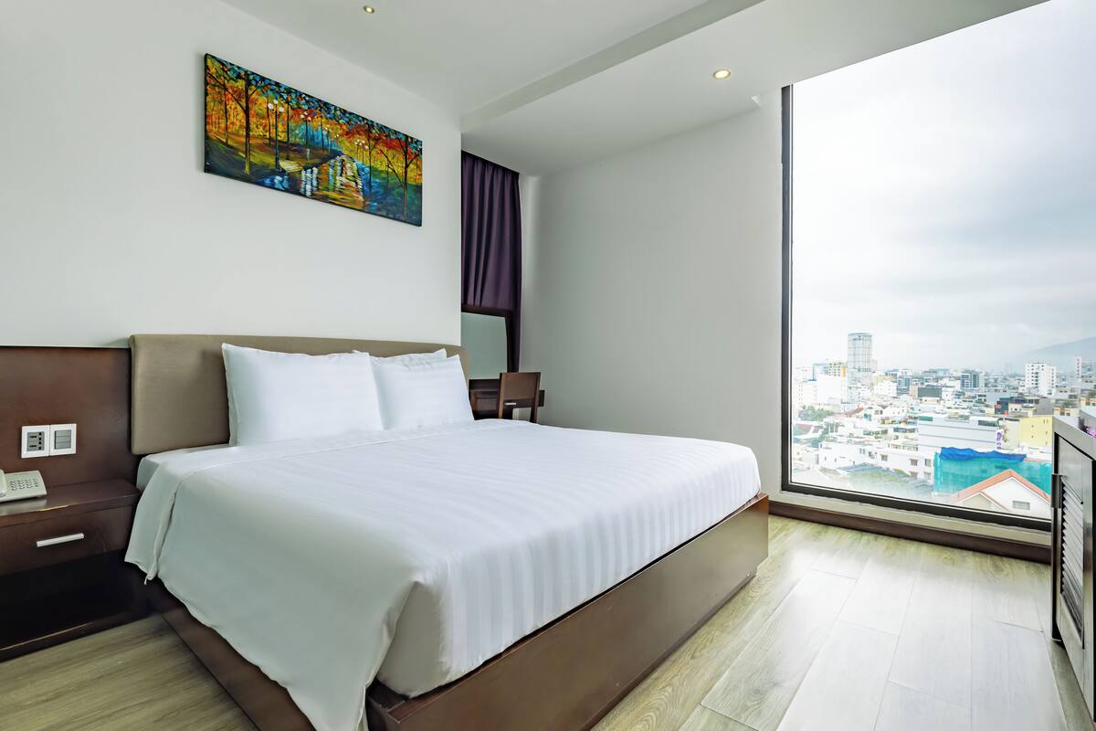 A modern hotel room with a large bed, a painting on the wall, a city view through the window, and a wooden floor.
