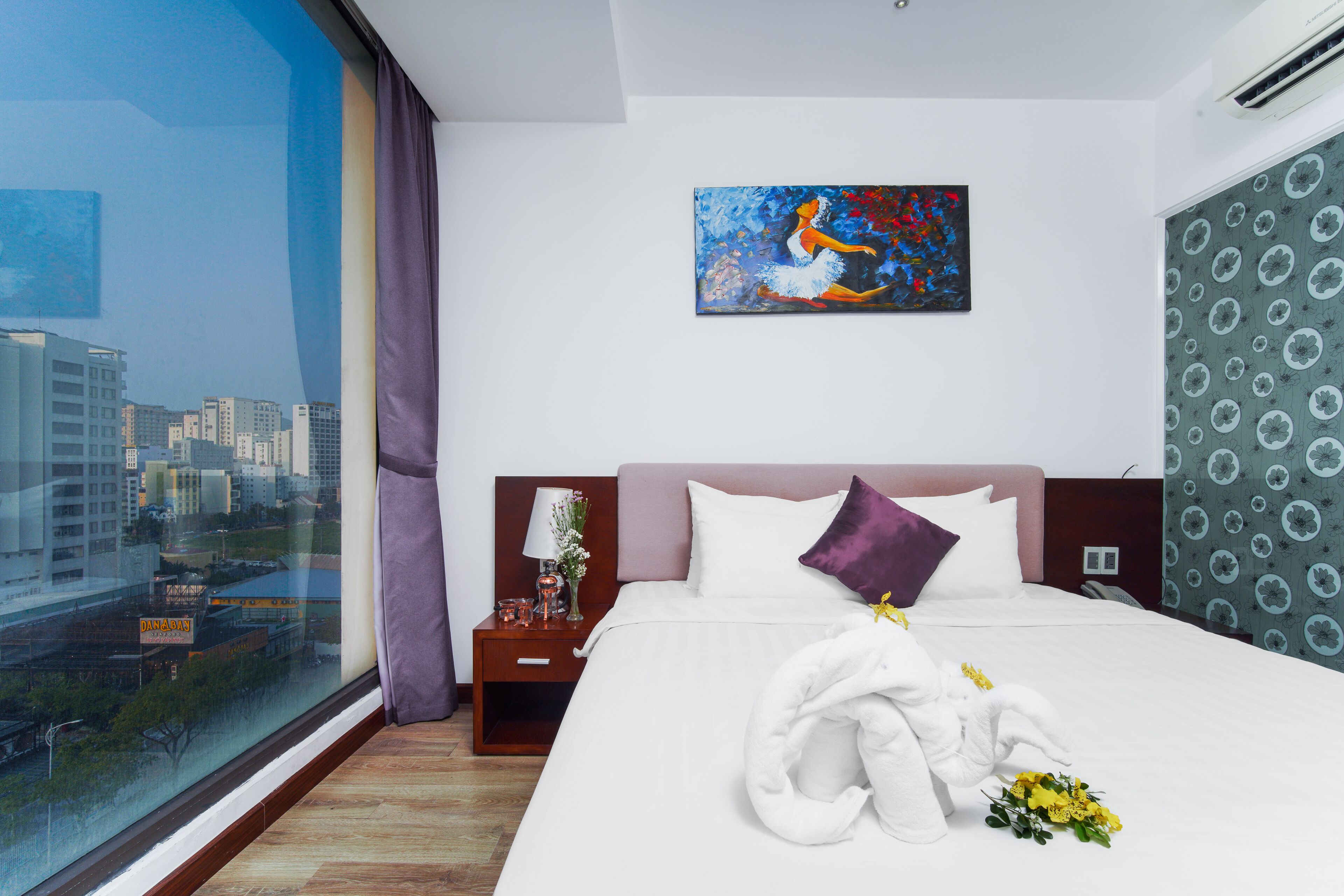 A hotel room with a large window, a bed with white linens, a purple pillow, and a painting on the wall.