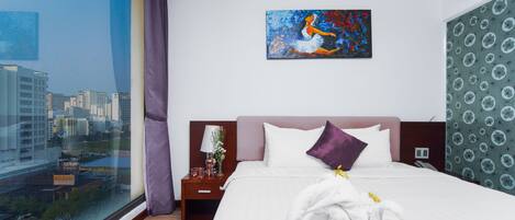 A hotel room with a large window, a bed with white linens, a purple pillow, and a painting on the wall.