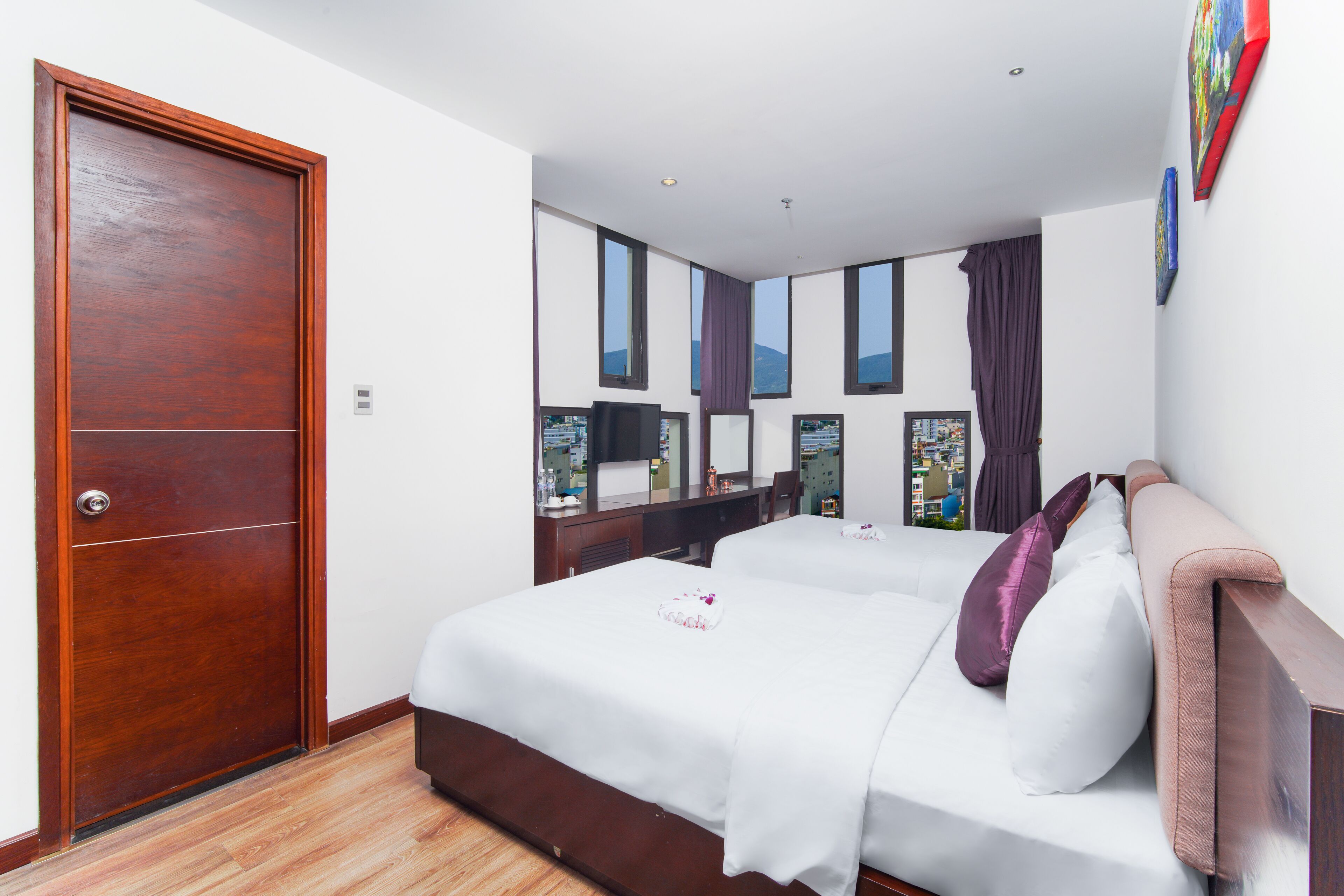 A hotel room with a wooden door, a bed with white bedding and a pink pillow, a desk with a computer, and a large window with a view.
