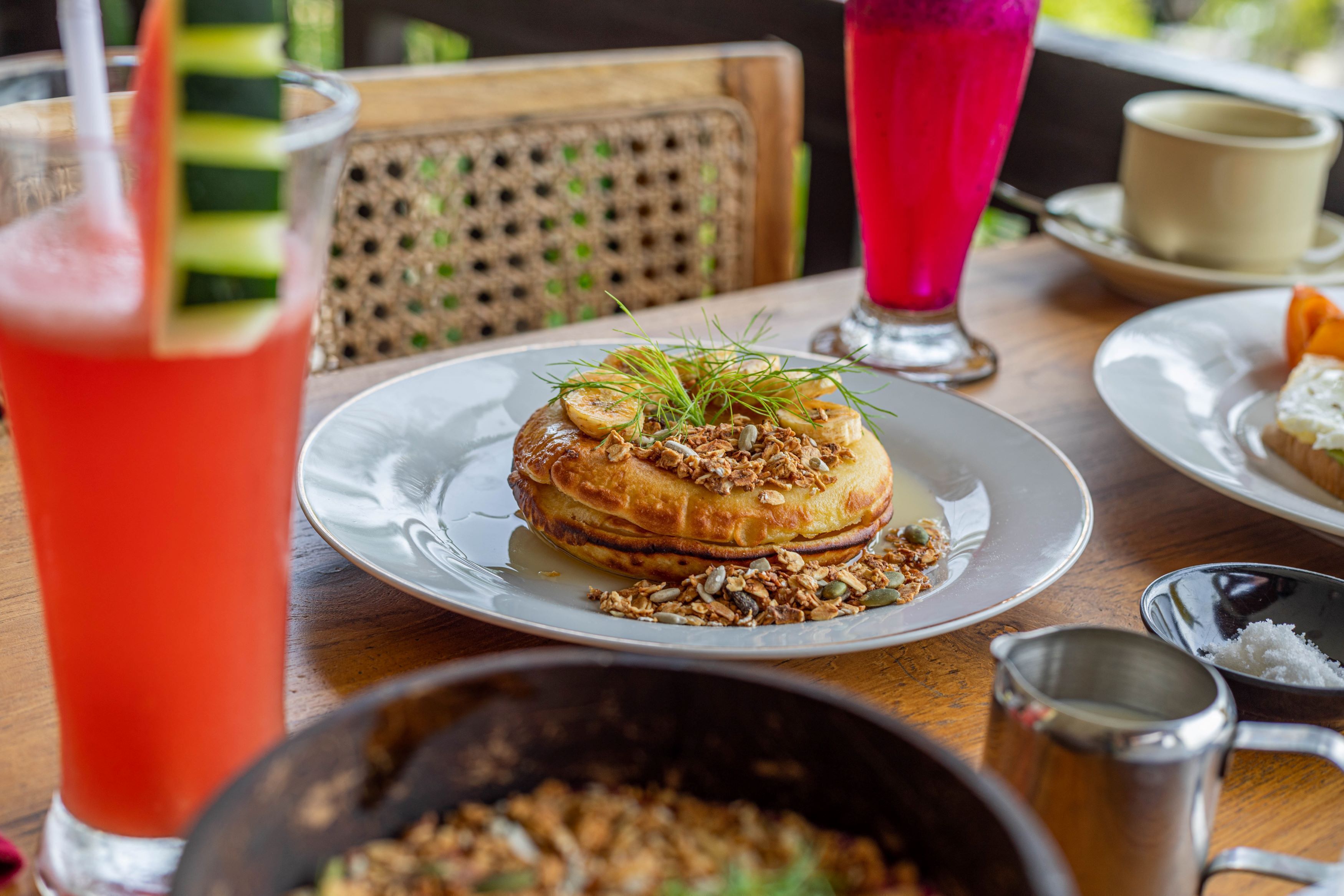 A table with a plate of pancakes topped with fruit and granola, a glass of red drink, and a pitcher of water.