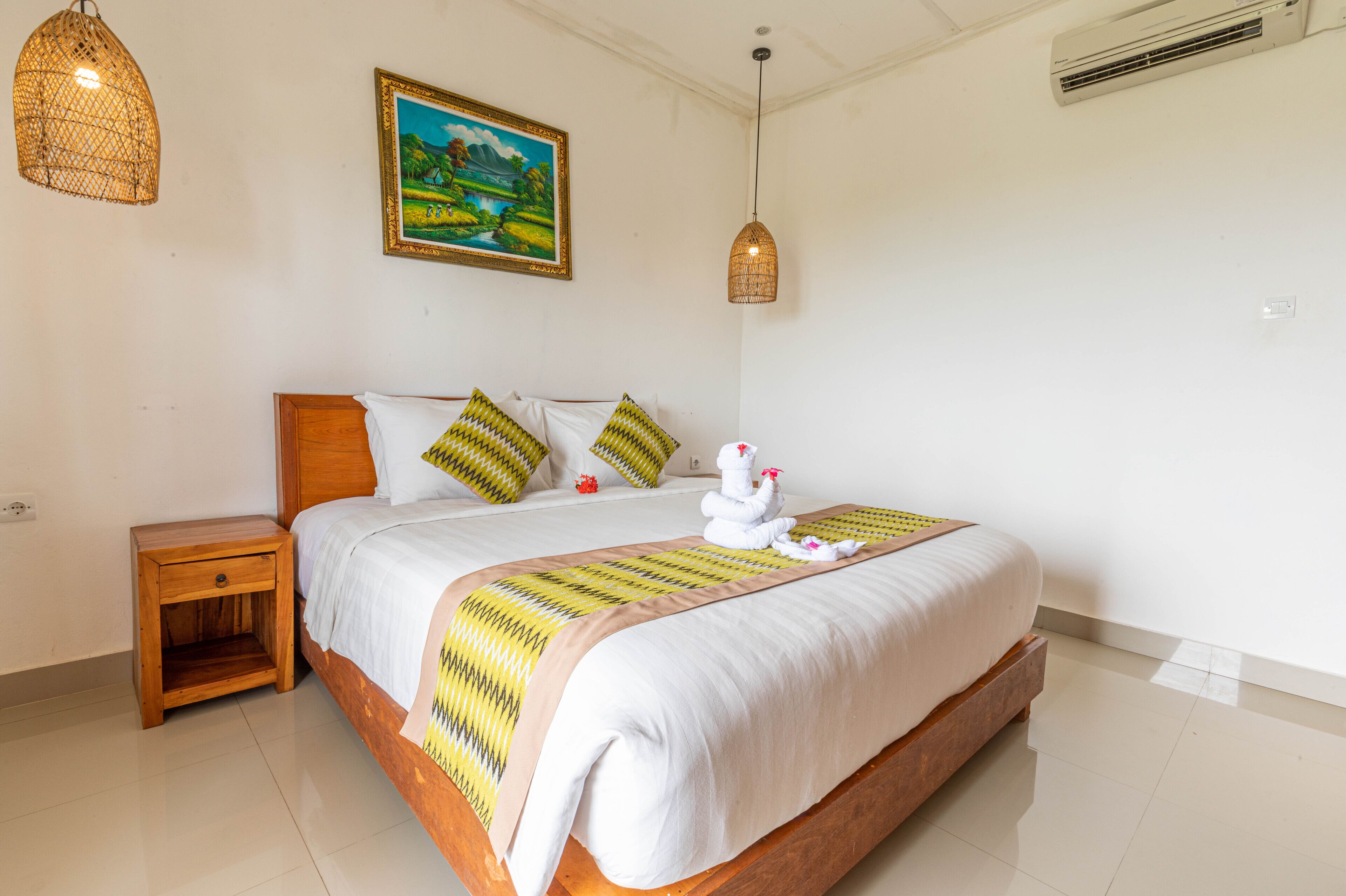 A neatly made bed with a white duvet and a decorative pillow, a wooden nightstand, a wall-mounted air conditioner, and a hanging light fixture.