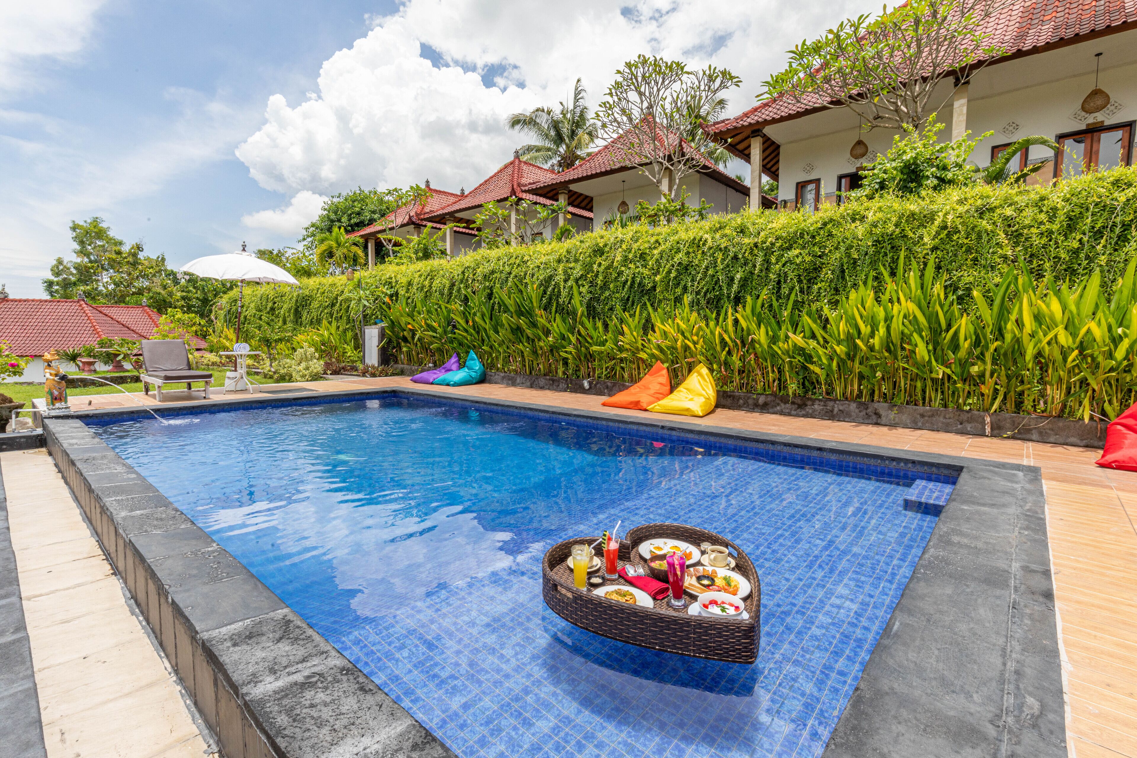 A swimming pool with a floating tray of food, surrounded by a well-maintained hedge and traditional-style buildings.
