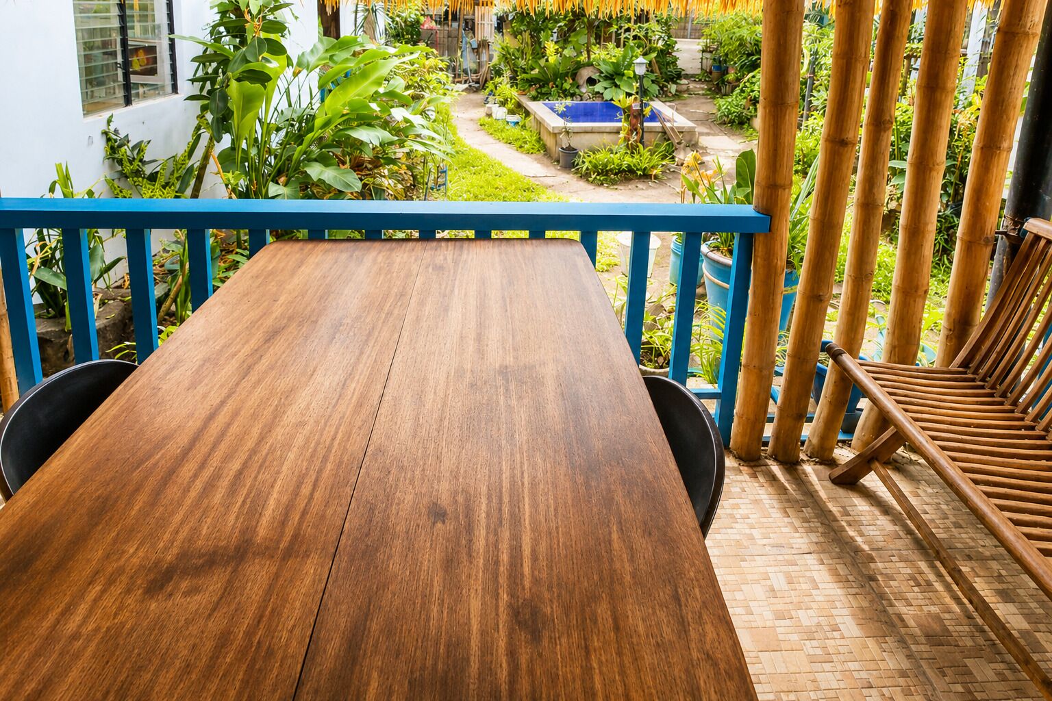 Una mesa de madera con sillas en un balcón con baranda azul, con vistas a un jardín con plantas diversas y un elemento acuático azul.