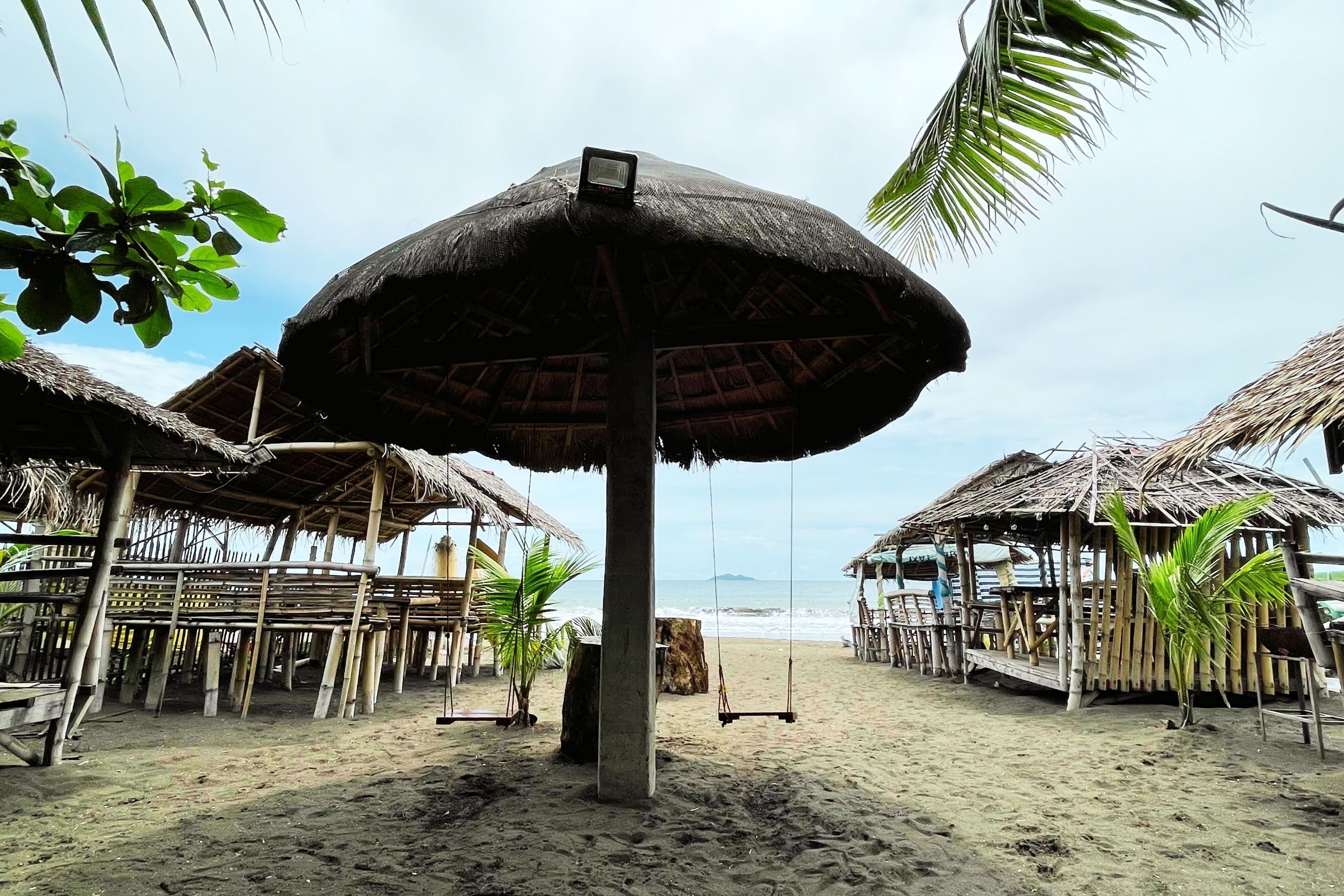 Een strand met constructies met rieten daken, waaronder een centrale parasol en schommels, en een houten omheining.