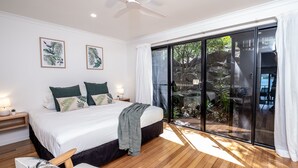Ein Schlafzimmer mit einem großen Bett, Holzboden, Deckenventilator und einer Glastür, die auf einen Balkon mit Pflanzen führt.