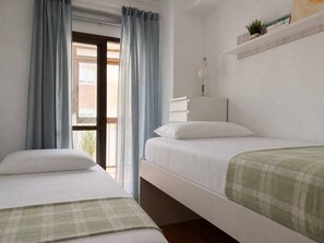 Un dormitorio con dos camas, una ventana con cortinas, una mesita de noche con lámpara, y una estantería con libros y una planta.