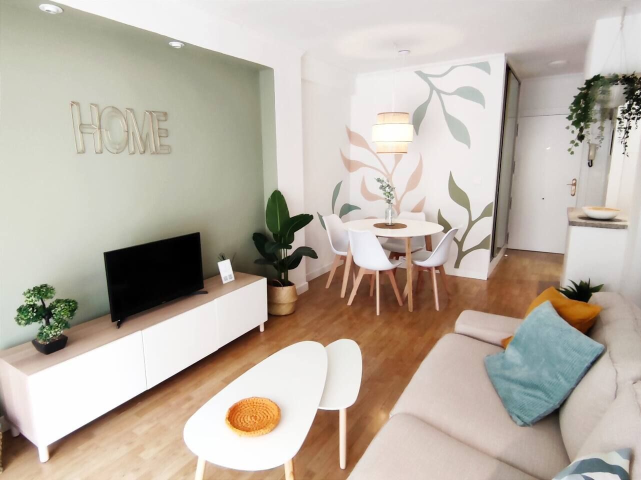 Una sala de estar moderna con un televisor de pantalla plana, un sofá blanco, una zona de comedor con mesa y sillas, y una pared con la palabra 'HOME' sobre una planta decorativa.