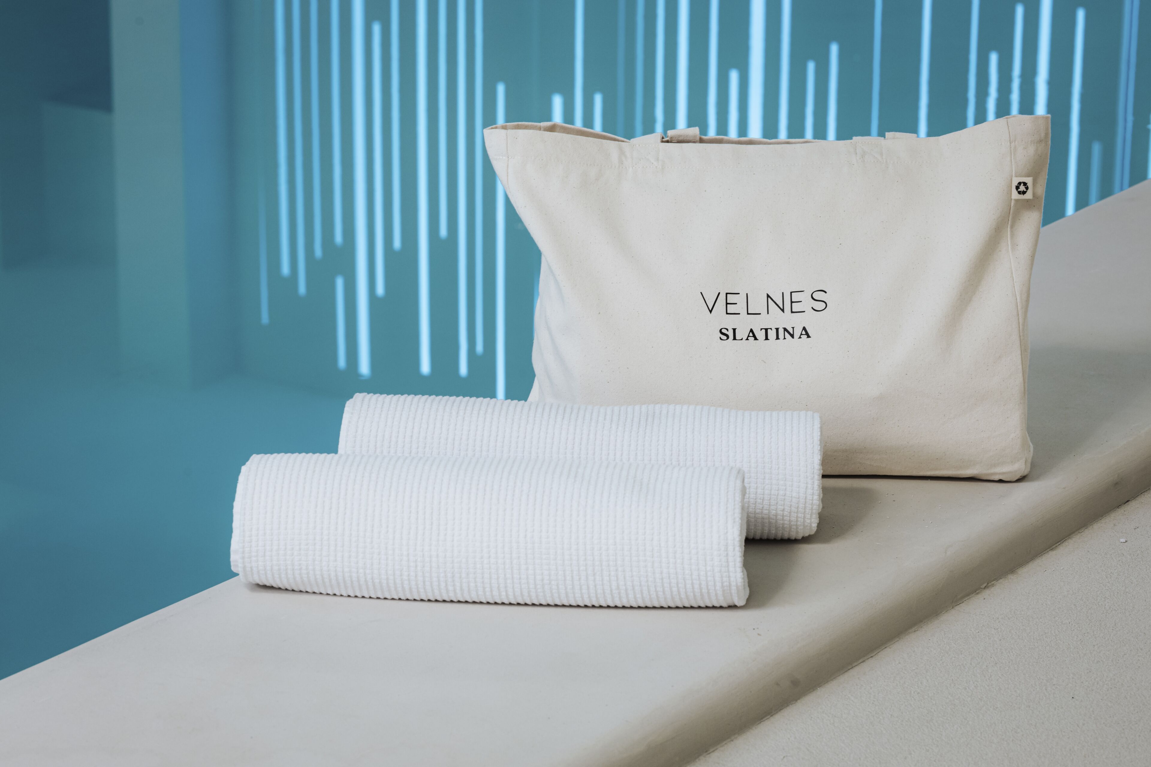 Toalha bege e almofada bege com o texto "VELNES SLATINA" sobre uma superfície branca com fundo azul.