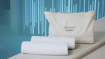 Toalha bege e almofada bege com o texto "VELNES SLATINA" sobre uma superfície branca com fundo azul.