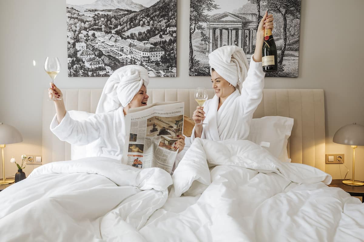 Two people in white bathrobes and towels, sitting on a bed with white bedding, holding wine glasses. They are reading a magazine. The room has a beige headboard and two bedside tables with lamps.