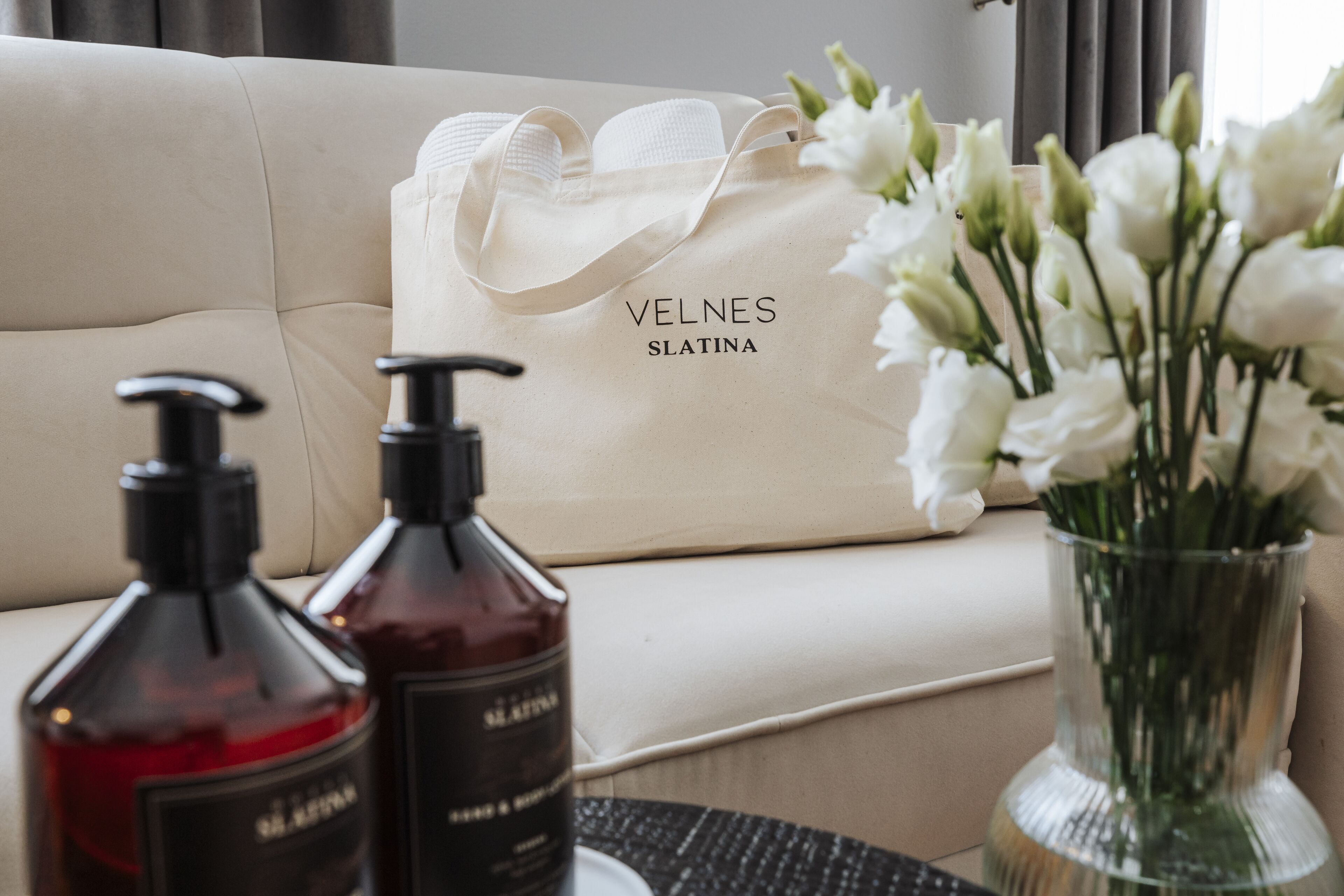 Un sofá beige con un bolso blanco Velnes Slatina y dos botellas de perfume sobre una mesa.