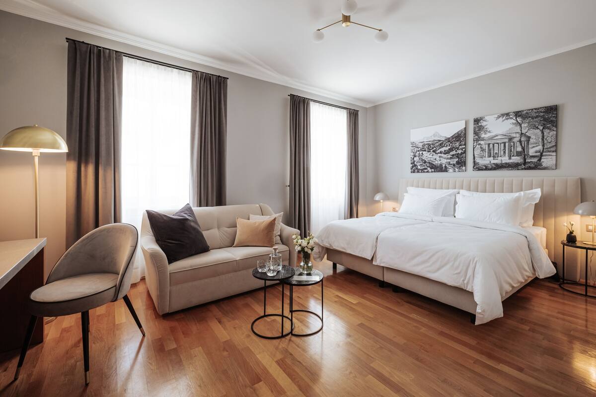 A modern hotel room with a bed, a sofa, a chair, a small table with a vase of flowers, and artwork on the wall.