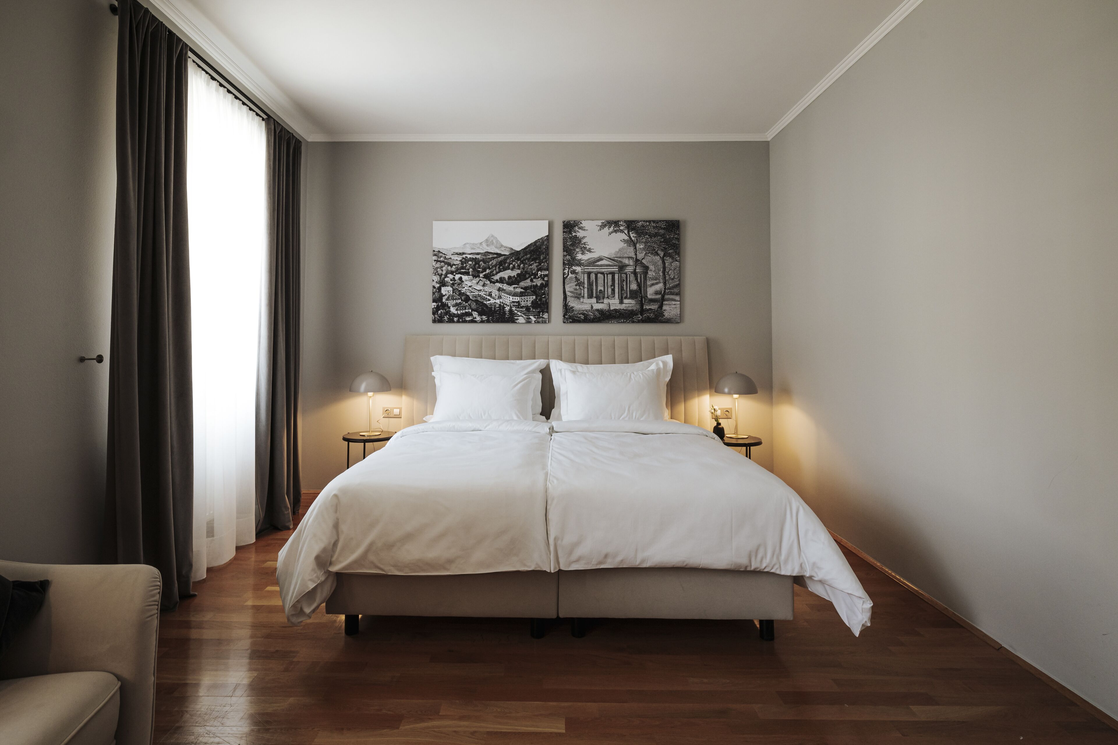 Een netjes opgemaakt bed met witte lakens, twee bedlampjes en twee aan de muur hangende schilderijen.