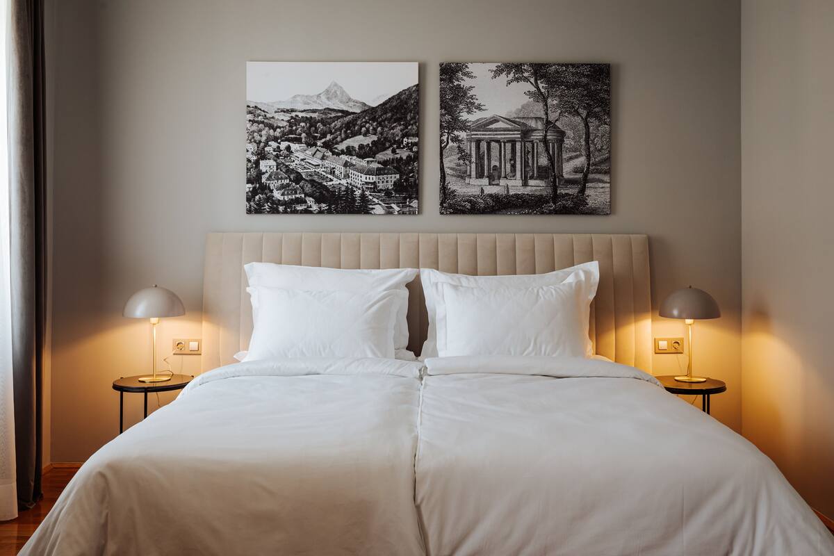 A neatly made bed with two white pillows and two bedside lamps. There are two black and white framed pictures on the wall above the bed.