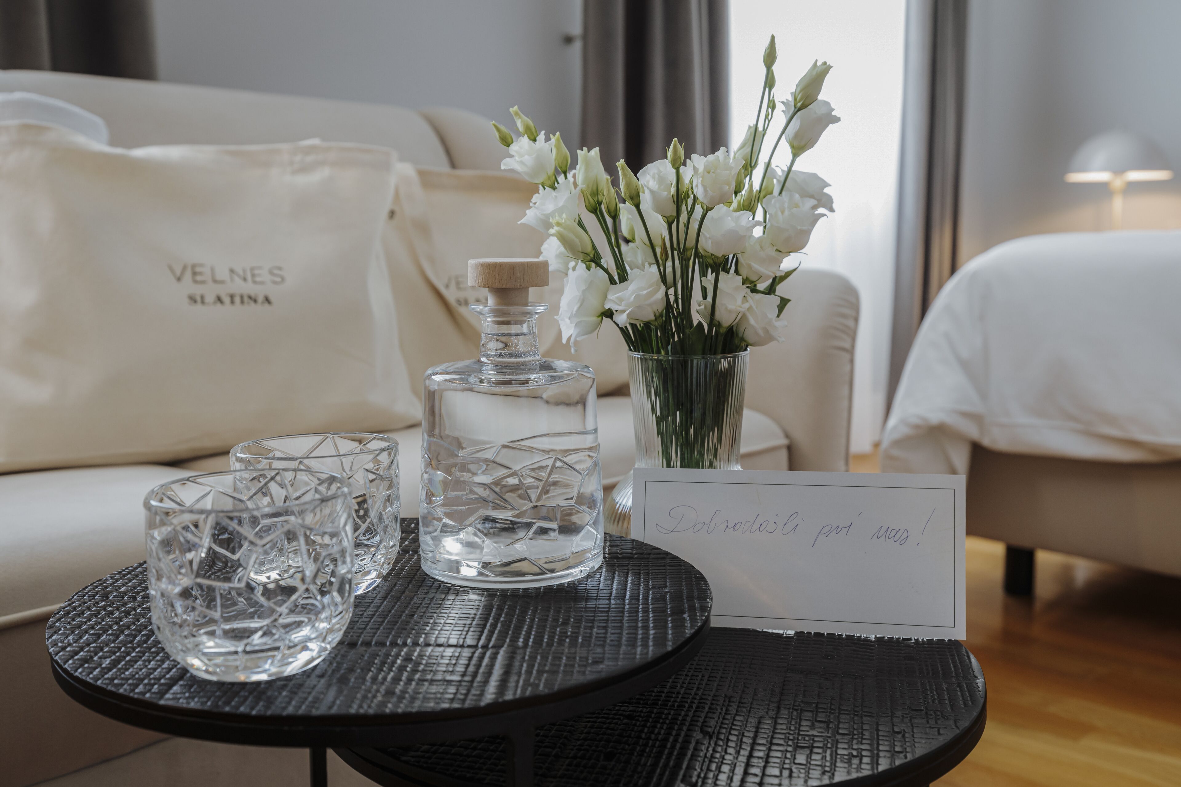 A table with a glass decanter, two glasses, a vase with white flowers, and a sign with text.