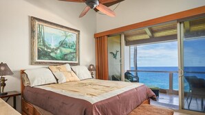 Une chambre à coucher avec un lit, un tableau, un ventilateur de plafond, une table de chevet et une vue sur l’océan par une porte-fenêtre coulissante.