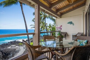 A covered outdoor seating area with wicker furniture and a glass table, overlooking a beach with palm trees and the ocean. - Aloha Condos, Keauhou Kona Surf and Racquet Club, Condo 2-302, Oceanfront, AC (Kailua Kona)