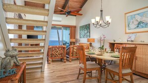 Un coin repas avec une table en bois, des chaises et un lustre. On y trouve un escalier avec des rampes en bois et un ventilateur de plafond.