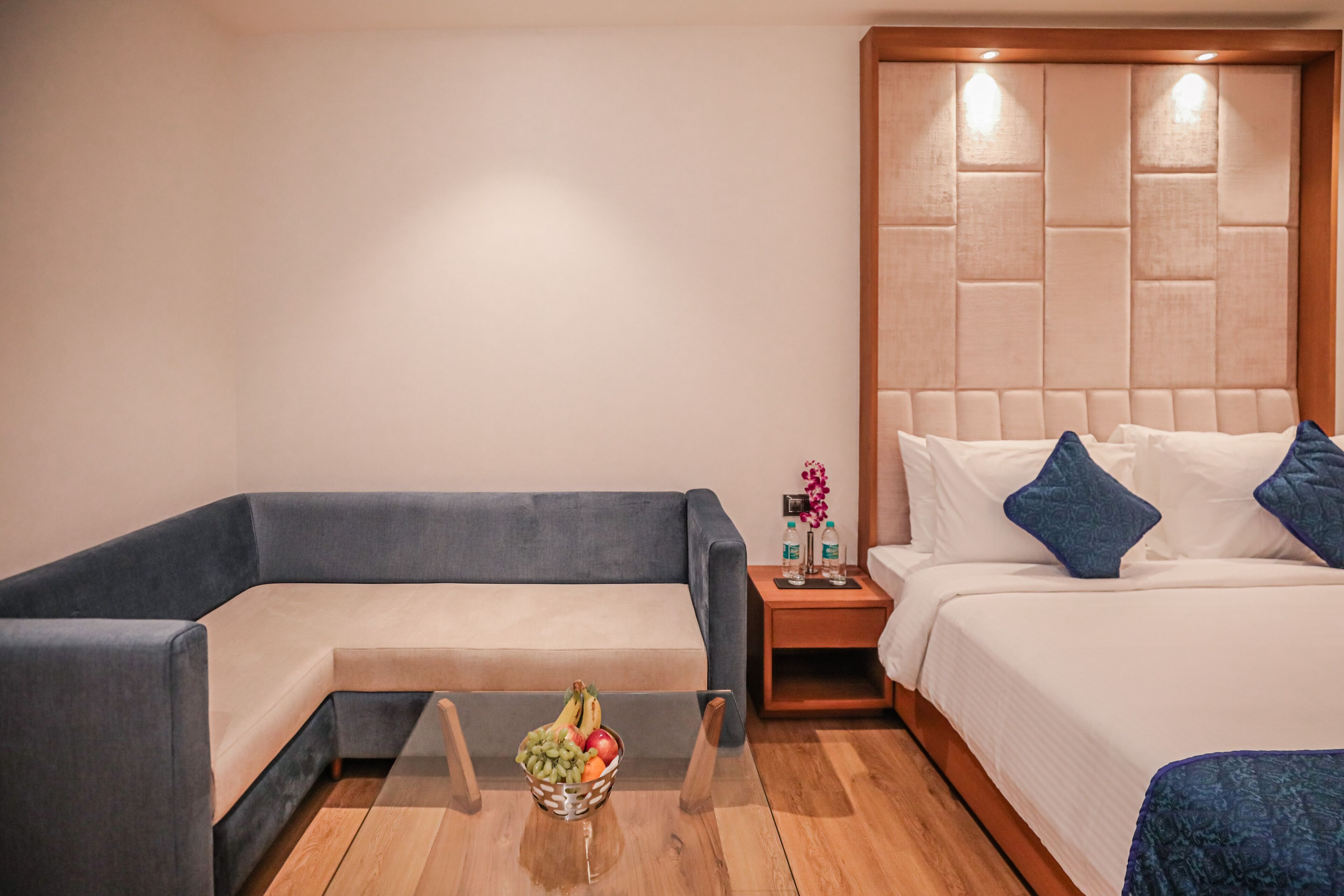 Une chambre d’hôtel moderne avec un canapé gris, une table basse en verre sur laquelle sont disposés des fruits, une table de chevet en bois avec des bouteilles d’eau, et un lit avec des oreillers blancs et bleus.