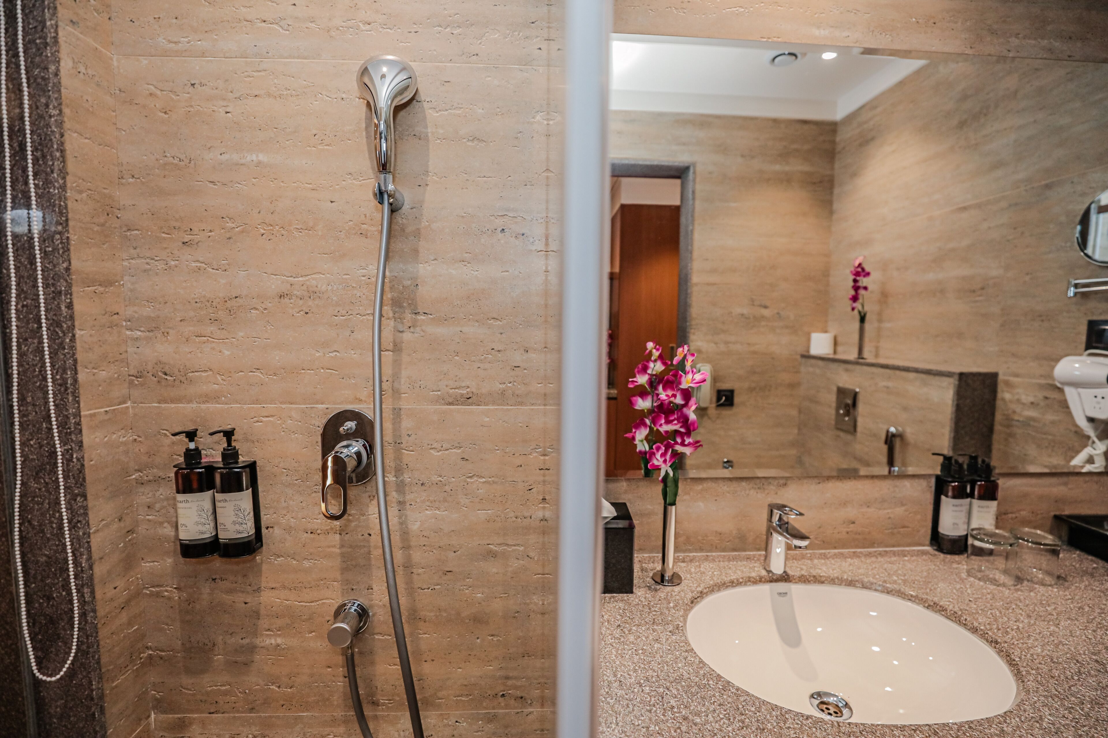 A modern bathroom with a beige marble wall, a white sink, and a shower with a handheld showerhead.